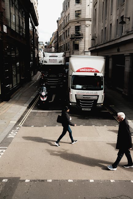 A street scene showing a white moving van with the logo 'uniserve' parked at the curb on a narrow urban road, with a second larger truck positioned behind it. The van's rear door is open, indicating loading or unloading activity related to house removals or furniture transport. A black motorcycle is parked on the left side of the van. In the foreground, two pedestrians are crossing the street; one a woman carrying a black bag in her right hand and walking briskly, the other an older man with grey hair walking in the opposite direction. The street is flanked by multi-storey buildings with large windows and fire escape balconies, and the scene is lit by natural daylight, suggesting a typical city environment suitable for professional relocation services such as those offered by Hounslow Man and Van.