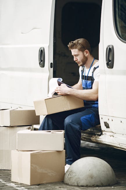 A young man with short brown hair and a beard, dressed in a blue and white striped t-shirt and blue overalls, is sitting inside the open sliding door of a white van. He is holding a cardboard box on his lap and appears to be reading or checking a document. Surrounding him on the ground are several additional cardboard boxes of various sizes, some stacked and others placed individually. The van's interior is visible behind him, showing a dark space where goods or packing materials might be stored during the house relocation process. The scene takes place outdoors on a paved surface, with a grey, rounded bollard in the foreground. This image represents aspects of furniture transport, packing, and loading processes involved in professional removals, as performed by services such as Hounslow Man and Van, which specialise in house removals and moving logistics.