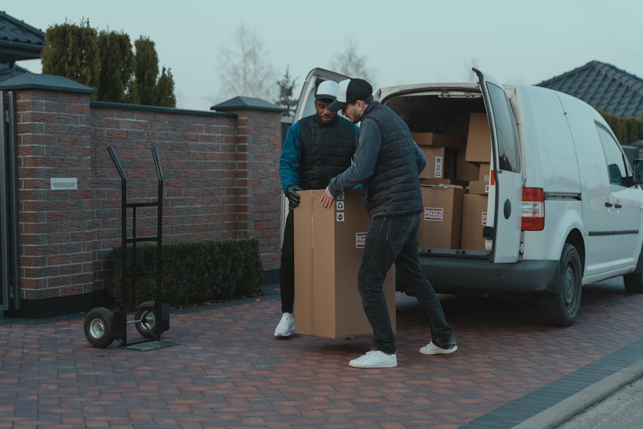 Two men are engaged in packing and loading a cardboard moving box into the back of a white van parked on a brick-paved driveway outside a residential property. One man, wearing a black quilted vest over a blue long-sleeve shirt, is assisting by holding the box, while the other, dressed in a dark hooded jacket and black pants, is carefully guiding it into the vehicle. An empty hand truck with black handles and two wheels is positioned nearby on the driveway, ready for use in furniture transport. Inside the van, several other boxes and packaging materials are visible, indicating an ongoing home relocation process. The setting is outdoors during daytime with overcast sky, surrounded by a brick wall and residential buildings with tiled roofs in the background. This scene exemplifies professional house removals and packing services offered by Hounslow Man and Van, supporting efficient and careful furniture transport and moving logistics.