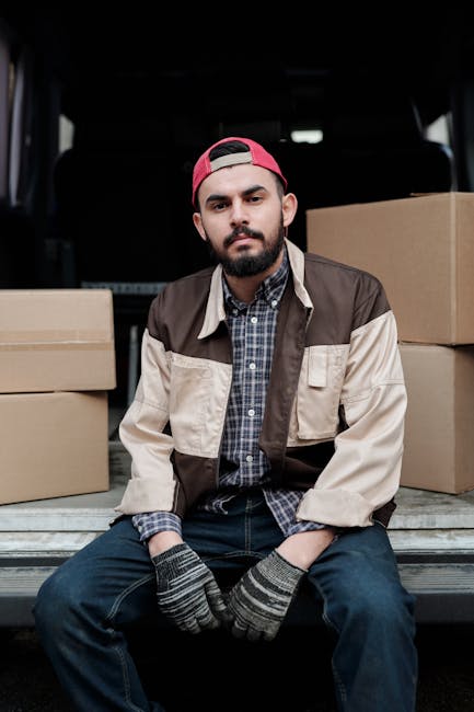 A young man with a beard, wearing a red cap backwards, a checked shirt, and a beige and brown jacket, is sitting on the edge of a van's open trunk during a home relocation. Surrounding him are several cardboard boxes of varying sizes, some stacked and others placed on the floor of the vehicle, all sealed with packing tape. The interior of the van is visible behind him, and he is wearing grey knitted gloves. The scene takes place outdoors on a paved surface, with natural lighting illuminating the loading process. The man appears to be taking a break during furniture transport or packaging and moving activities, which are part of a professional removals service offered by Hounslow Man and Van, as indicated by their website. The focus is on the loading process of house items, with the boxes prepared for transport as part of the furniture and household packing involved in residential relocation.
