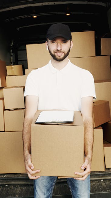 A man and a woman are inside a room with wooden flooring, surrounded by multiple cardboard moving boxes of various sizes, some labeled with handwritten text such as 'BOOKS'. The woman is seated on the floor, holding an open book, while the man is leaning over a box, looking at her. Both are dressed casually, with the woman wearing light-colored clothing and the man in a grey hoodie. In the background, a large window with curtains brings natural light into the space, and additional boxes are stacked against the wall. The scene appears to be part of a home relocation process involving packing and organizing items for a move, with the process of loading and transporting belongings supported by professional moving services, such as those offered by Hounslow Man and Van.