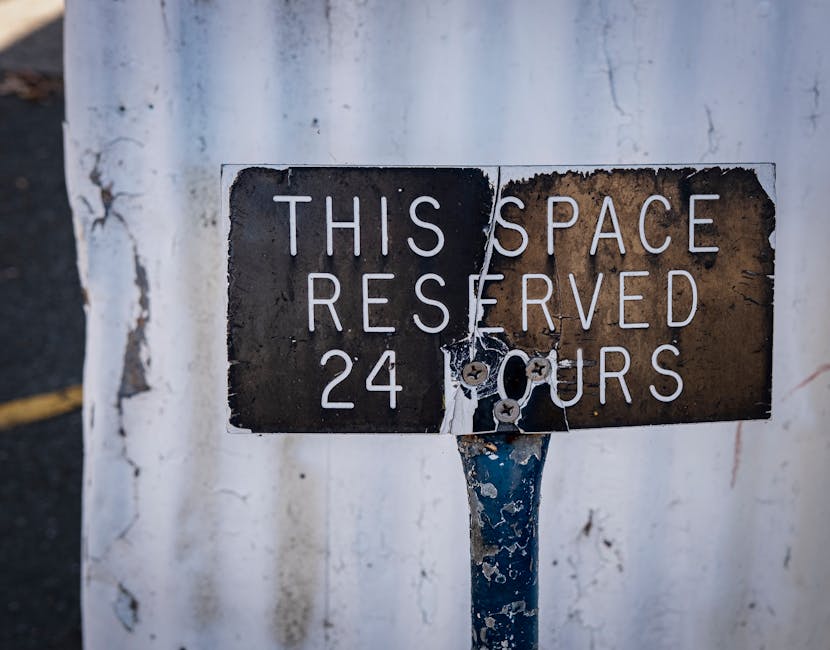 Close-up of a weathered metal parking restriction sign mounted on a metal pole, with chipped and peeling black and white paint, indicating that the space is reserved 24 hours. The sign is attached to a white wall or barrier that shows signs of wear, including cracks and peeling paint. This image relates to house removals and moving services, where parking permissions and street restrictions are important during the loading and unloading process. [COMPANY_NAME], a professional removal service, adapts to such parking rules to ensure smooth furniture transport and packing during a home relocation, especially in urban areas like Hounslow. The environment suggests an outdoor setting with natural light, emphasizing the importance of understanding street regulations in moving logistics.