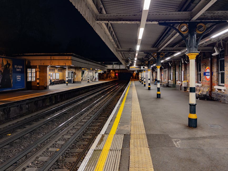 The image shows Hounslow West railway station platform in the evening, illuminated by overhead lighting, with multiple sets of train tracks running through the station. The platform is empty except for a small seating area with benches, and a person sitting near a building on the left side. The station architecture includes brick walls, metal columns with decorative yellow and black striped bands, and shelter roofing supported by metal beams. There are tactile paving strips along the edge of the platform for accessibility, and a sign indicating the station's name on the side of a structure. The setting suggests a quiet period during a home relocation process involving careful planning and transport logistics, possibly by professional removals services like Hounslow Man and Van, who may facilitate safe and efficient furniture transport and packing during house moves.