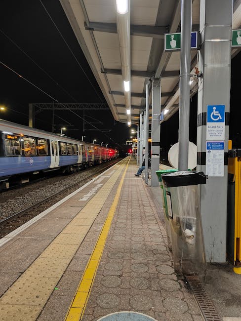 A view of multiple modern yellow and gray commuter trains parked on several railway tracks at Hounslow West station, with some trains in focus near the foreground and others extending into the distance. The tracks are lined with metal rails, wooden sleepers, and gravel, and a tall fence runs alongside the right side of the tracks for safety. Overhead electrical wires and supporting poles are visible above the trains, indicating an electrified rail system. In the background, there are several multi-story residential and commercial buildings, with some trees and overcast sky further enhancing the urban setting. The scene captures the station's loading and parking area during daylight hours, providing a clear perspective on the rail infrastructure used for home relocation or furniture transportation in the Hounslow area, as managed by Hounslow Man and Van.