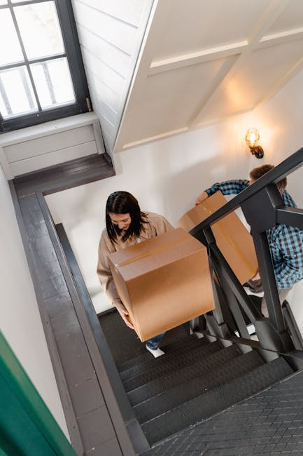 A woman with long dark hair, dressed in a beige top and blue jeans, is carrying a large cardboard box up a narrow staircase inside a home. The staircase has dark carpeting with a black metal handrail on the right side, and the walls are painted white. A second person, partially visible and wearing a blue and black checkered shirt, is behind her also holding a similar box. Natural light enters through a window at the top of the stairs, and a wall-mounted light fixture illuminates the space. The scene depicts the process of home relocation or furniture transport as part of a professional removal service offered by Hounslow Man and Van, who specialise in house removals and moving logistics.