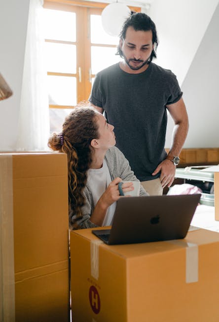 A woman with red hair and a white top, seated at a table with an open laptop, is surrounded by several cardboard boxes used for home relocation, with one directly in front of her. She is engaging in c