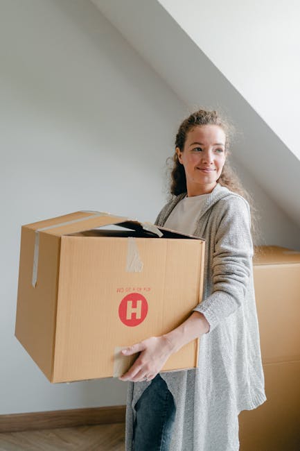 A woman with red hair and a white top, seated at a table with an open laptop, is surrounded by several cardboard boxes used for home relocation, with one directly in front of her. She is engaging in c