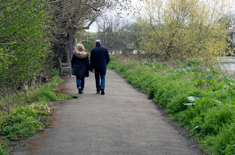 A middle-aged couple, wearing dark jackets and jeans, walk hand-in-hand along a narrow paved pathway lined with green shrubs and trees on either side. The woman has a fur-lined collar on her coat, and the man has short gray hair. They are heading away from the camera, towards a bench placed under a large tree with sprawling branches and fresh spring leaves. The pathway appears to be part of a peaceful park or riverside setting, with some buildings visible in the distance through the trees. The scene is illuminated by soft, natural daylight, suggesting an overcast or partly cloudy day. This outdoor environment, with its lush greenery and tranquil atmosphere, reflects typical surroundings for residential relocations or moving scenarios, where careful packing, loading, and transportation of furniture and boxes may take place nearby. Hounslow Man and Van offers services related to home relocation, including loading and transport, as part of comprehensive house removals.