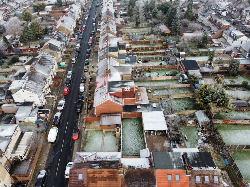 An aerial view of a residential street during daytime showing parked cars lining both sides of the road, with snow-dusted roofs and gardens. The houses are semi-detached and terraced, with enclosed backyards featuring various fencing, sheds, and garden furniture. Some properties have driveways, while others have small front gardens. A narrow alleyway runs parallel to the street, lined with trees and shadowed by the surrounding houses. This scene illustrates a typical UK suburban neighbourhood where home relocation and furniture transport might be coordinated by [COMPANY_NAME], with the street environment providing context for loading or unloading processes during same-day removals and moving services.