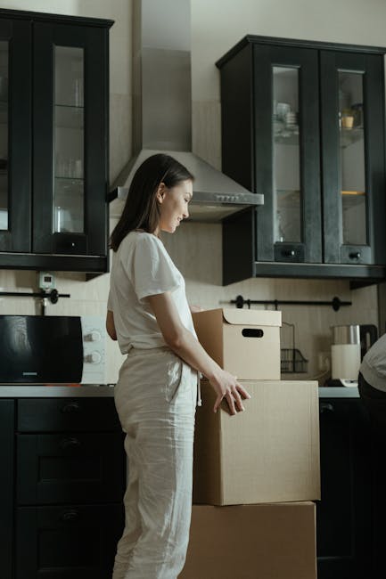 A woman with long dark hair, dressed in a white outfit, is standing inside a modern kitchen preparing to load or unpack cardboard boxes. She is positioned near dark-colored kitchen cabinets with glass doors, which contain dishes and glassware, and a stainless steel range hood is visible above the cooking area. The woman appears focused on her task, with her right hand resting on a medium-sized cardboard box, while another larger box is placed on a countertop or table in front of her. Various packing materials and appliances, including a black toaster and a white kettle, are seen on the countertop behind her. The environment is well-lit with natural or bright artificial light, emphasizing the clean and organised space. This scene illustrates home relocation or packing activities as part of a house move, which [COMPANY_NAME], Hounslow Man and Van, provides within the context of professional removal services, supporting seamless furniture transport and moving logistics.