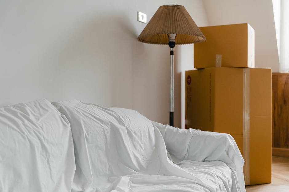 Interior view of a room with white walls and wooden flooring, showing several cardboard moving boxes stacked next to a standing floor lamp with a brown fabric shade. The boxes vary in size and are sealed with packing tape, with one partially obscured by the others. A wooden piece of furniture is visible in the background, and the boxes are positioned near a doorway or window, as indicated by the natural lighting. The scene suggests a typical house relocation process, with boxes prepared for moving. The company Hounslow Man and Van's removal services in Hounslow are relevant to this setting, reflecting packing and moving preparations for a home transfer, in line with the TW3 flat removals specialist service for Lampton Road moves.